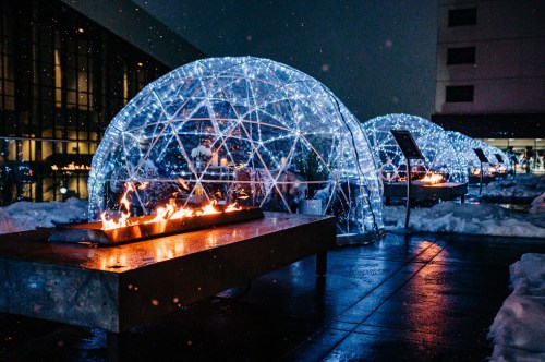 Illuminated igloos with fire pits, surrounded by snow, create a cozy atmosphere at night.