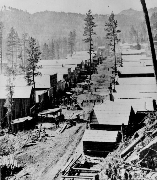 Historic black-and-white photo of a mining town with wooden buildings and trees in the background.