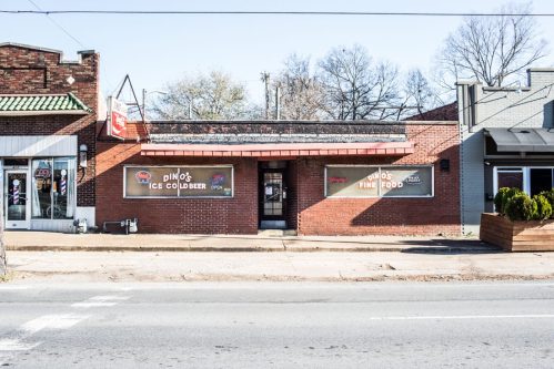A brick storefront featuring "Dino's Ice Cold Beer" and "Dino's Fine Food" signs, with a clear blue sky above.