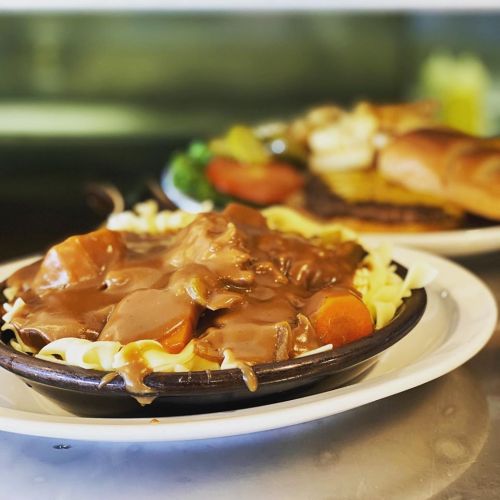 A plate of noodles topped with beef and gravy, accompanied by vegetables in the background.