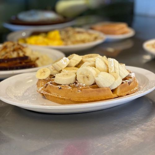 A plate of waffles topped with sliced bananas and powdered sugar, with other breakfast dishes in the background.