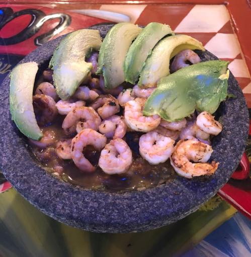 A stone bowl filled with shrimp and topped with slices of avocado. Colorful background with a checkered pattern.