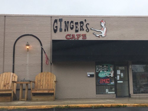 Exterior of Ginger's Cafe featuring a sign, large wooden chairs, and an open sign in the window.