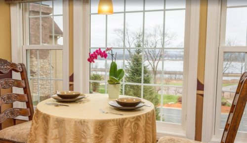 A cozy dining table set for two, with an orchid centerpiece and a view of a lake through large windows.