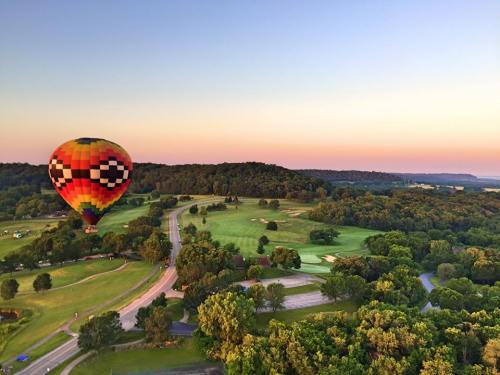 A colorful hot air balloon floats over a lush green landscape at sunset, with winding roads and trees below.