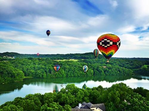 Colorful hot air balloons float over a lush green landscape and a serene lake under a partly cloudy sky.