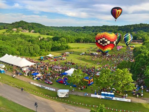 A vibrant outdoor festival with colorful hot air balloons, a large crowd, and various attractions on a green field.