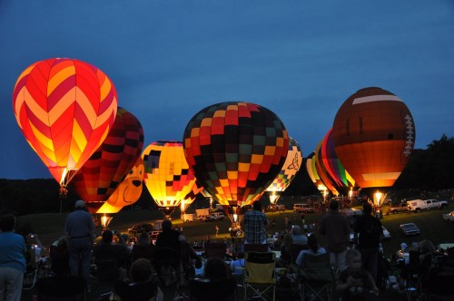 Colorful hot air balloons illuminated at night, with spectators seated on the ground enjoying the display.