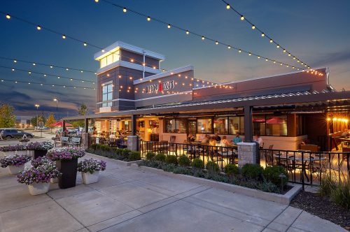 A modern restaurant with outdoor seating, string lights, and flower planters, set against a twilight sky.