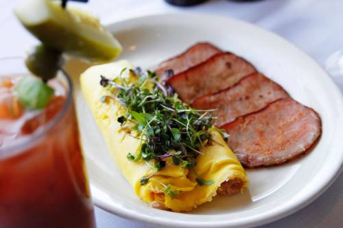 A plate featuring a rolled omelet with greens and slices of bacon, alongside a cocktail garnished with a pickle.