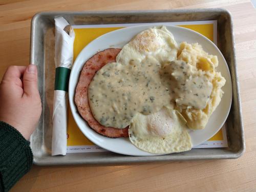 A plate with two fried eggs, ham, mashed potatoes, and gravy, served on a tray with a napkin and utensil.
