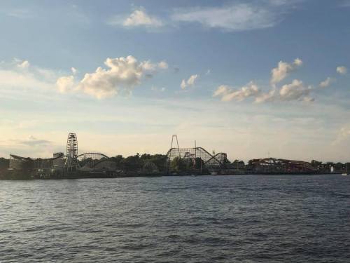 A scenic view of a waterfront amusement park with rides, including a Ferris wheel and roller coaster, under a blue sky.