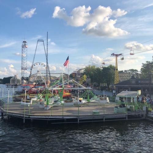 A vibrant amusement park on a waterfront, featuring rides, a Ferris wheel, and a zip line under a blue sky.