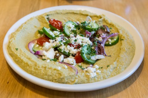 A plate of hummus topped with diced tomatoes, cucumbers, red onions, herbs, and crumbled feta cheese.