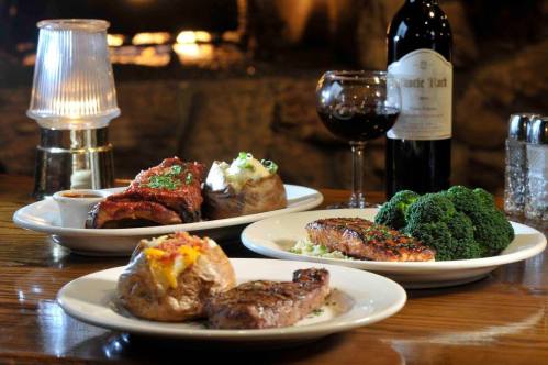 A table setting with three plates of gourmet food, a bottle of wine, and a glass of red wine, with a cozy background.