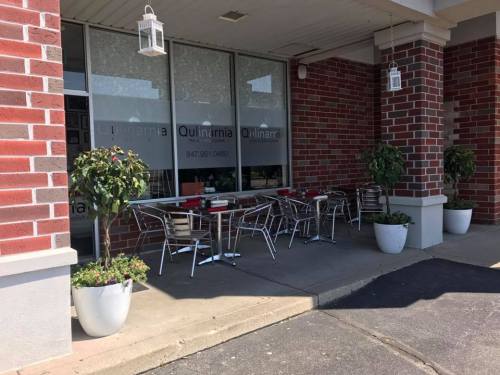 Outdoor seating area with metal tables and chairs, surrounded by potted plants, at a restaurant with large windows.