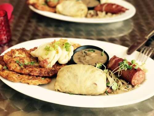 A plate of food featuring cabbage rolls, sausage, potato pancakes, and a small bowl of gravy, garnished with green onions.