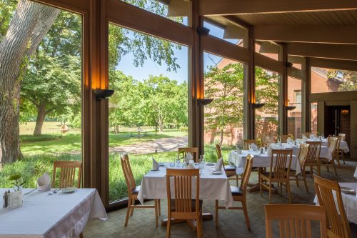 Bright dining area with wooden chairs and tables, large windows showcasing a green outdoor view.