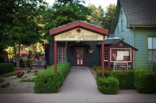 A charming restaurant entrance with a red sign reading "Red Geranium," surrounded by greenery and flowers.