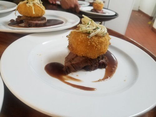 A plate featuring sliced beef topped with a golden, crispy ball and drizzled with sauce, garnished with herbs.