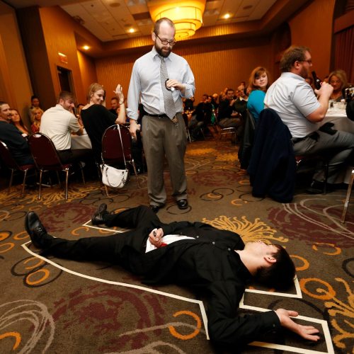 A man in a suit lies on the floor while another man stands nearby, speaking to a crowd at a banquet event.