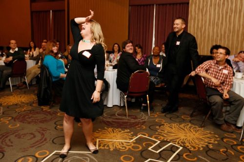 A woman in a black dress playfully dances in front of a group at a social event, with tables and attendees in the background.