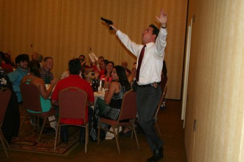 A man in a suit holds a gun while raising his arms, entertaining a seated audience at a banquet.