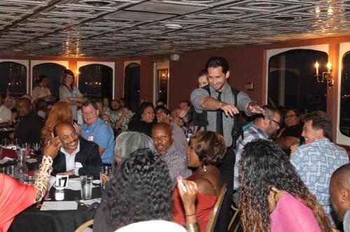 A lively dinner event with a performer engaging a laughing audience at tables, creating a fun atmosphere.