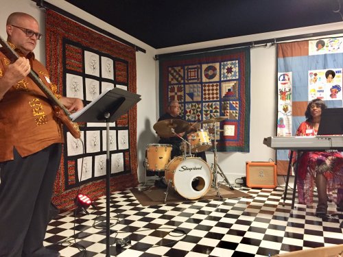 A jazz trio performs in a colorful room with quilts and artwork, featuring a bassist, drummer, and keyboardist.