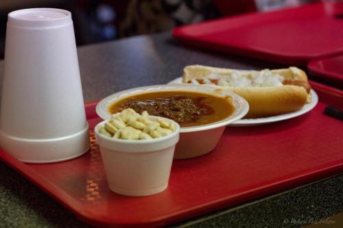 A tray with a hot dog topped with onions, a bowl of chili, and a cup of crackers, alongside a white drink cup.