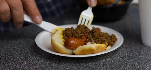 A hand holds a fork over a hot dog topped with chili on a paper plate.