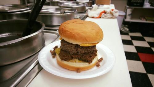 A large hamburger with two beef patties, cheese, and pickles on a white plate in a kitchen setting.