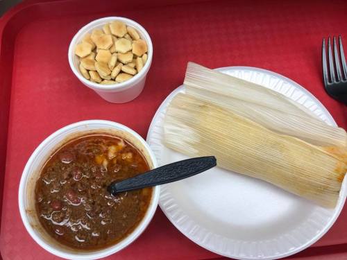 A red tray with a bowl of chili, a tamale on a plate, and a cup of crackers.