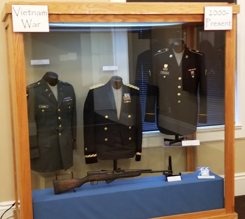 Display case featuring military uniforms from the Vietnam War and 2000-Present, with a rifle in the center.