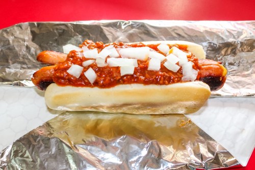 A hot dog topped with chili and diced onions, served in a soft bun and wrapped in foil.