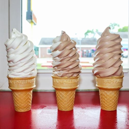 Three soft-serve ice cream cones in waffle cups: one vanilla, one chocolate, and one swirl, displayed by a window.