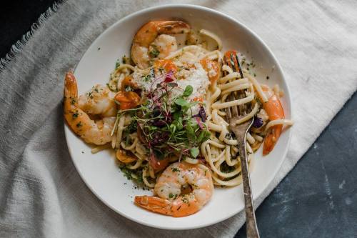A plate of shrimp pasta garnished with herbs and microgreens, served with a fork on a textured cloth background.