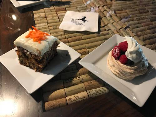 A slice of carrot cake with cream cheese frosting and a pavlova topped with berries and whipped cream on white plates.
