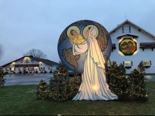 A large illuminated nativity scene featuring Mary and baby Jesus, with a decorated building and Christmas trees in the background.