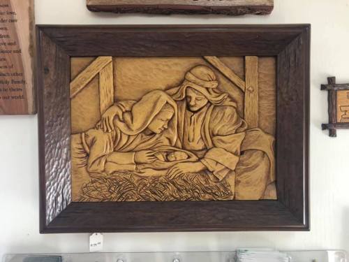 A carved wooden relief depicting two women admiring a newborn in a manger, framed and displayed on a wall.