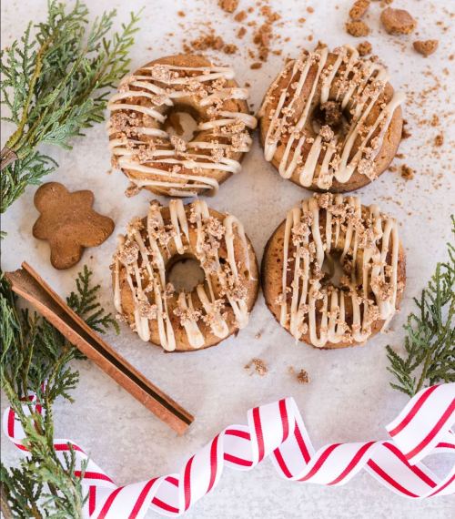Four decorated donuts with icing and crumbs, surrounded by greenery, a gingerbread cookie, and a red-striped ribbon.
