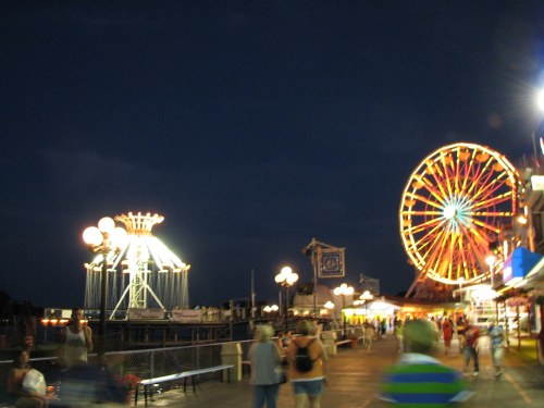 A vibrant amusement park at night, featuring a Ferris wheel and a spinning ride, with people walking along the boardwalk.