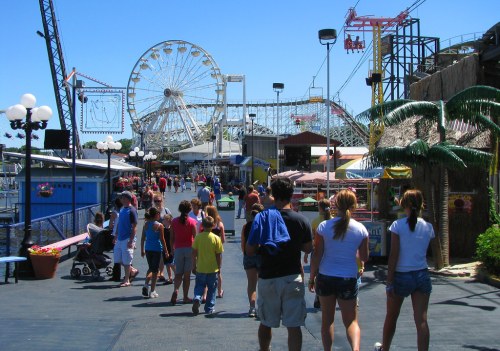 A busy amusement park boardwalk with a ferris wheel, rides, and people walking in a sunny atmosphere.