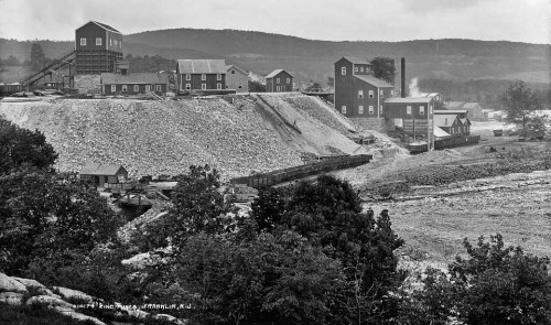Historic black-and-white photo of a mining site with buildings on a hillside and a train near a river.