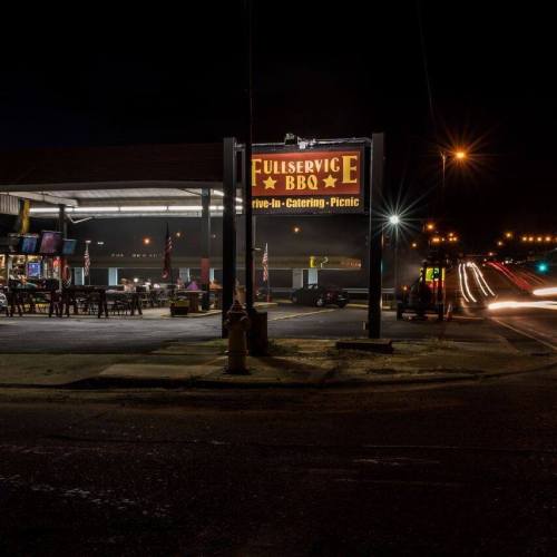A nighttime view of Full Service BBQ, featuring a bright sign and outdoor seating, with traffic lights in the background.