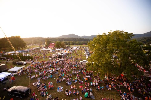A large crowd gathers on a grassy field at sunset, with tents and cars in the background and mountains in the distance.