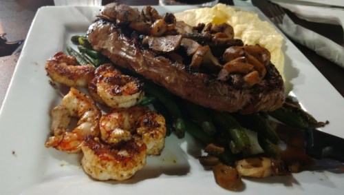 A plate featuring a grilled steak topped with mushrooms, shrimp, green beans, and creamy mashed potatoes.