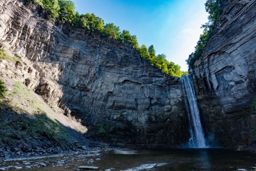 A tall waterfall cascades down rocky cliffs, surrounded by lush green trees under a bright blue sky.
