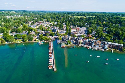 Aerial view of a lakeside town with a marina, boats, and lush green surroundings on a sunny day.