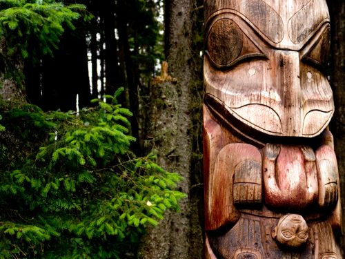 A wooden totem pole with intricate carvings stands among lush green trees in a forest setting.